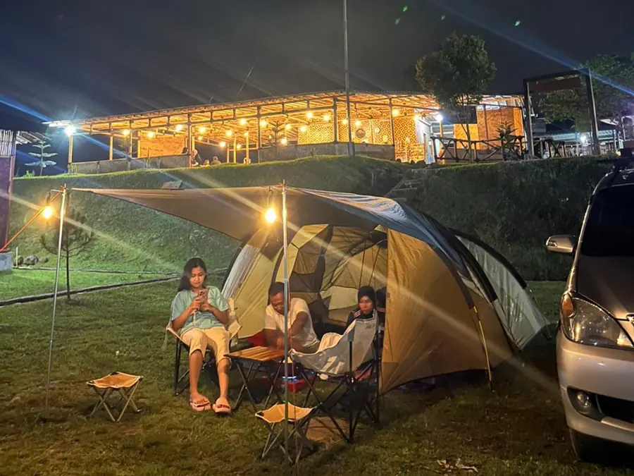 Resto Sisi Cai terang berhias lampu di malam hari, dengan keluarga bersantai di depan tenda